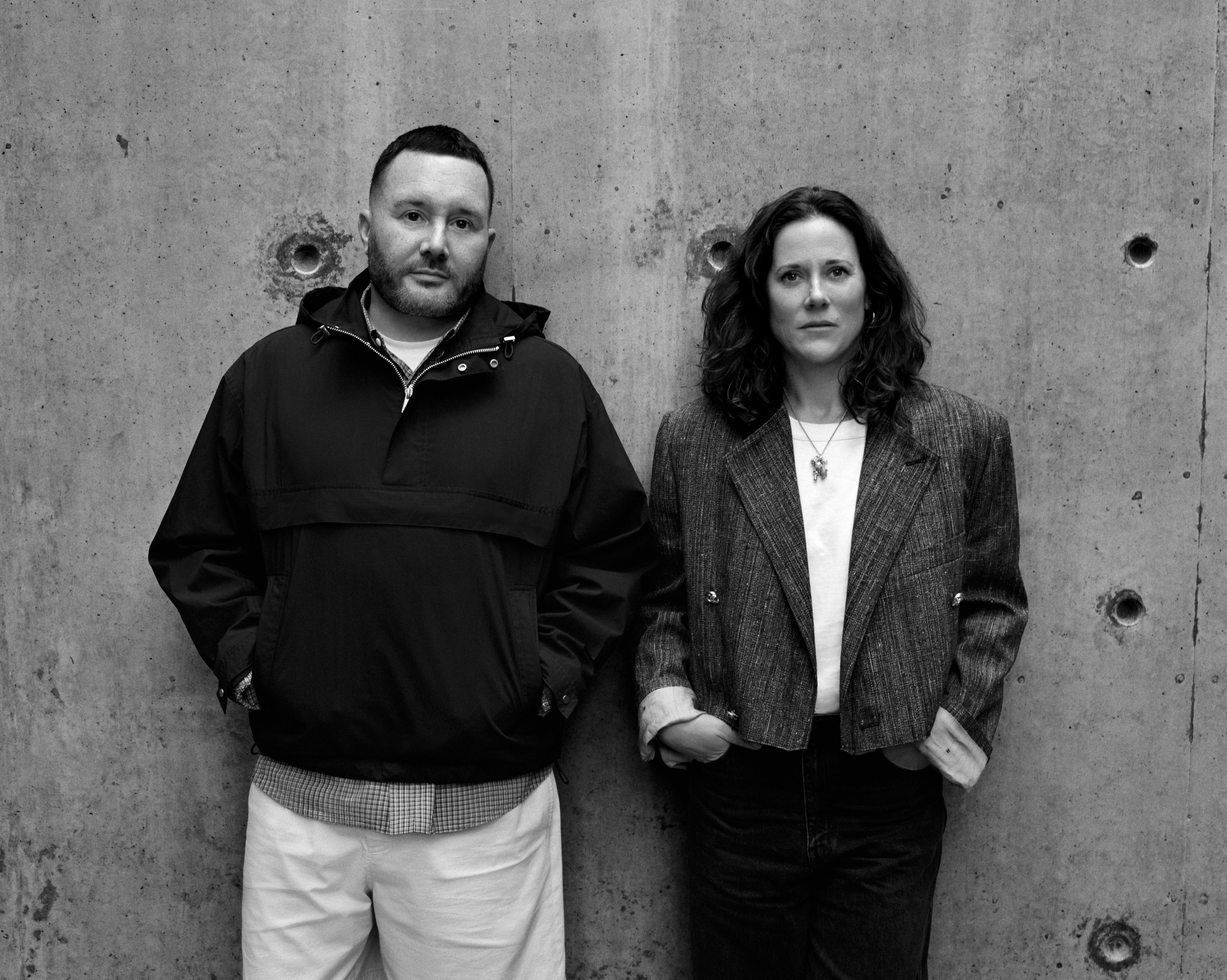 This black and white photograph shows two individuals standing against a concrete wall. The man on the left is wearing a black jacket and white pants, with his hands in his pockets. The woman on the right is wearing a gray blazer over a white shirt and dark-colored jeans. Both individuals are looking directly at the ca