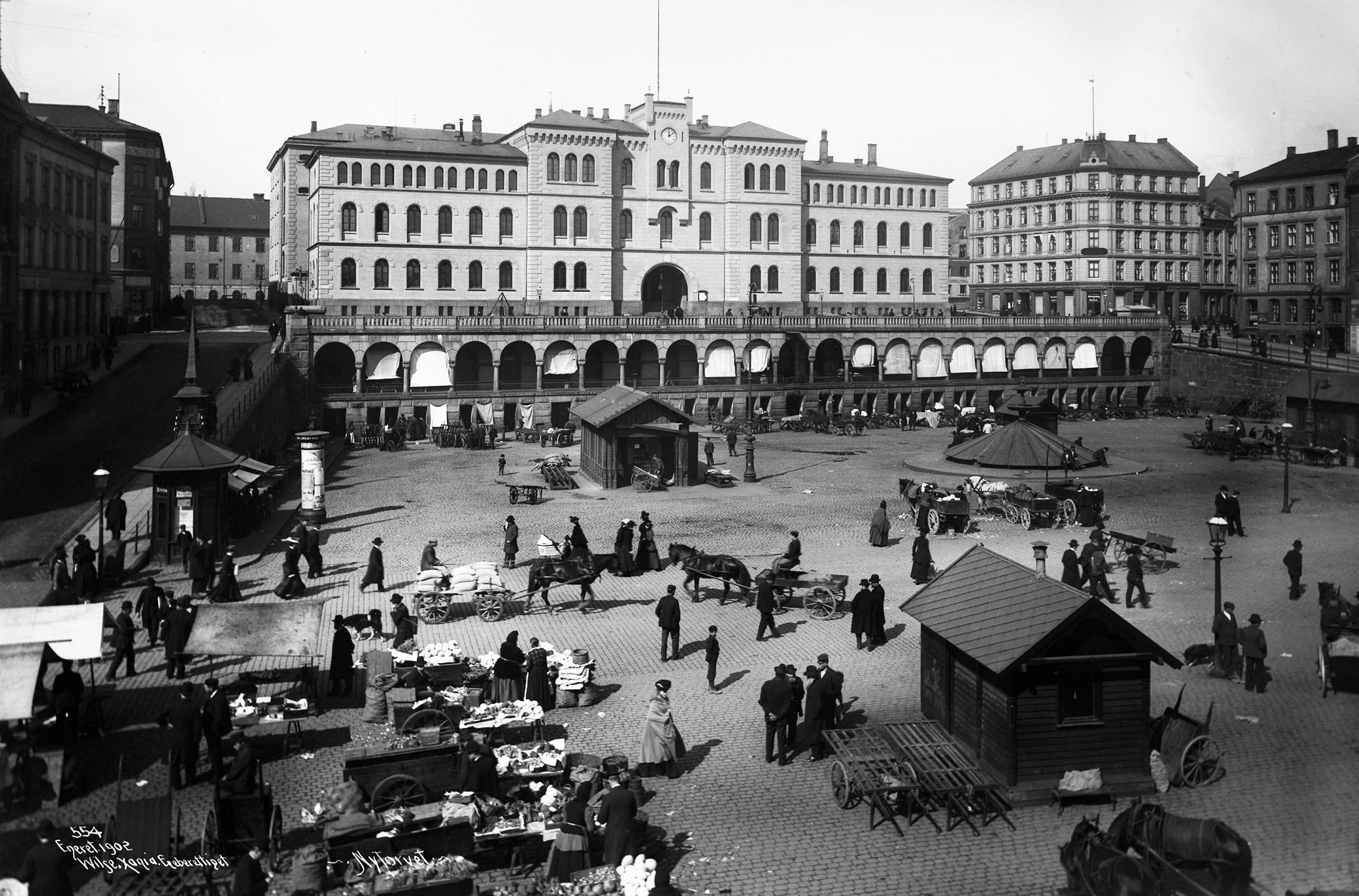 NYTORGET 1902: «Ute på torvet ved den store vekten fant man kjøtthandlerne med sine lass av dyreskrotter.»     Haakon B. Nielson. Se godt etter så sees både vekt og dyreskrotter ved det bakerste lille skuret. Til høyre for det sees også den tildekka fontena Hedvig Camilla lekte ved som barn. I 1942 ble den fjerna og angivelig dumpa i havne    bassenget. En kopi er seinere oppført. 