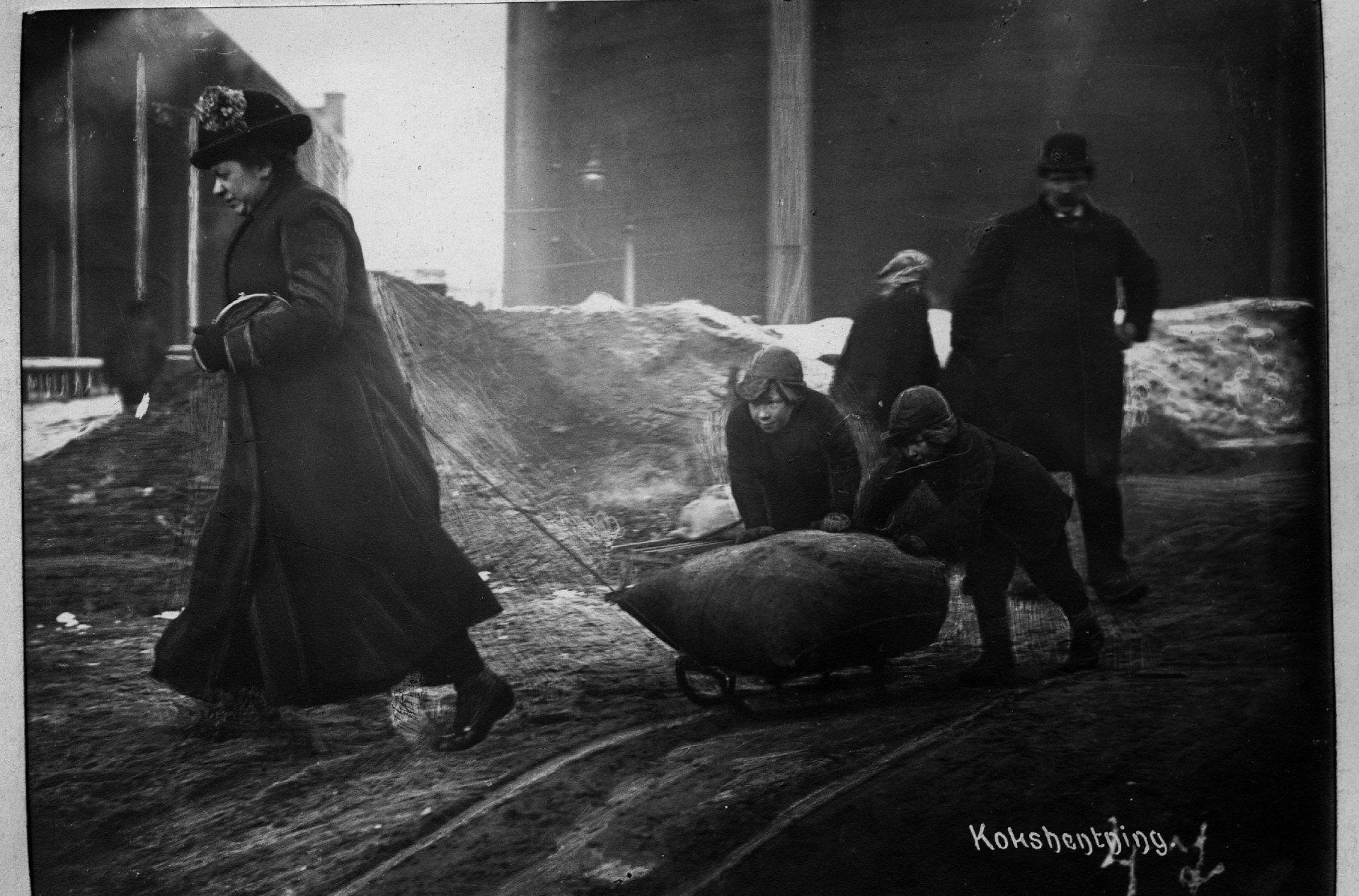 Koksen var billig, men støvete og tung. En kvinne med kåpe, hatt og veske drar en kjelke med en kokssekk fra Ankertorget til Hammersborg mens to gutter skyver og mannen står og ser på. Vinteren 1919. Foto: Narve Skarpmoen/Oslo byarkiv