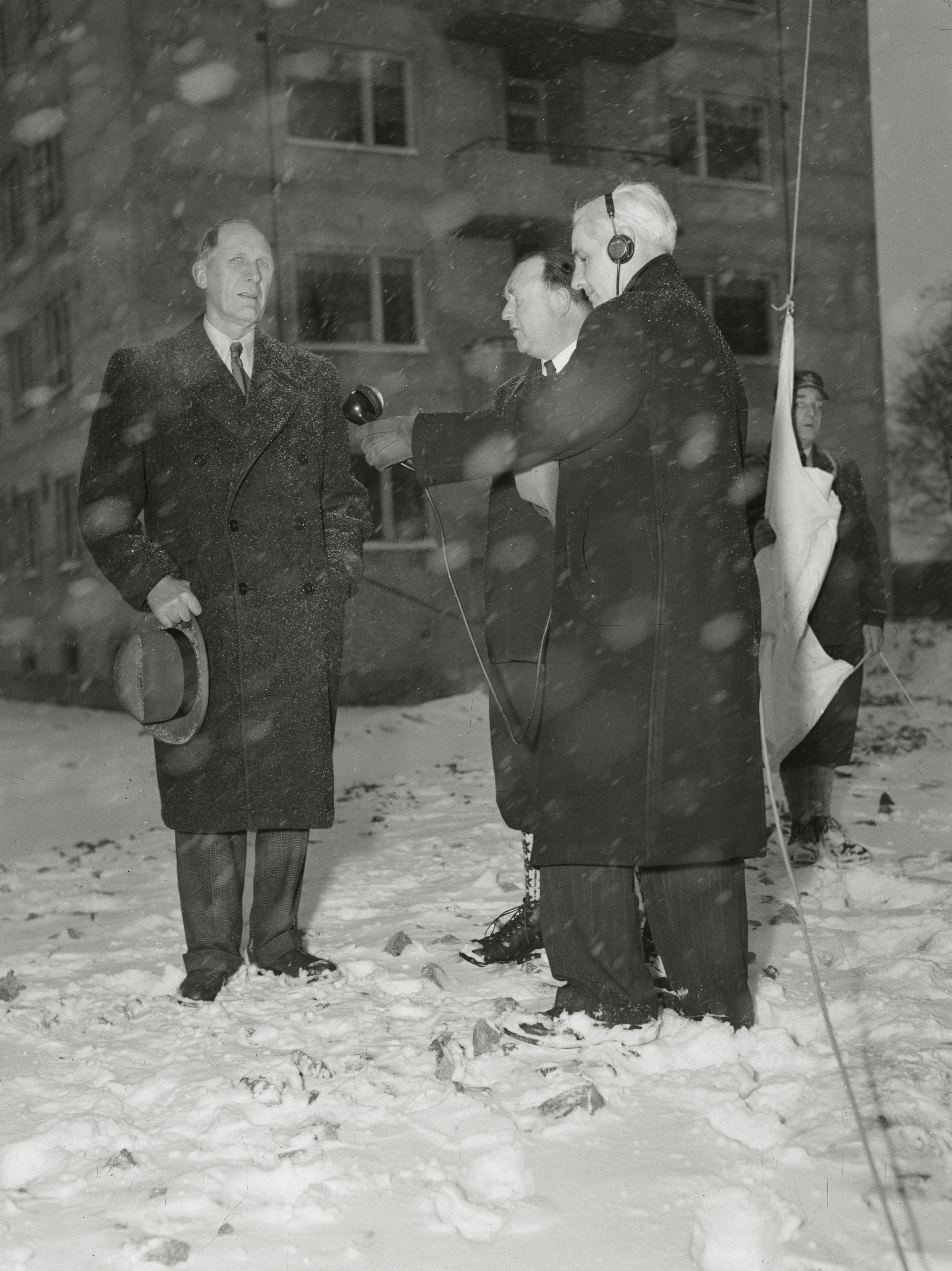 Været var helt krise under Vinter-OL i 1952: Isete og lite snø, og tett sludd eller tåke. Her holder formannen i vinterlekenes organisasjonskomite, O. Ditlev Simonsen jr., en kort tale i olympialeiren på Ullevål, som blir kringkastet. Med mikrofonen står NRKs nyhetsredaktør Karl C. Lyche, og i midten innkvarteringssjef Aksel W. Floer. Til høyre er olympiaflagget klart til å heises.