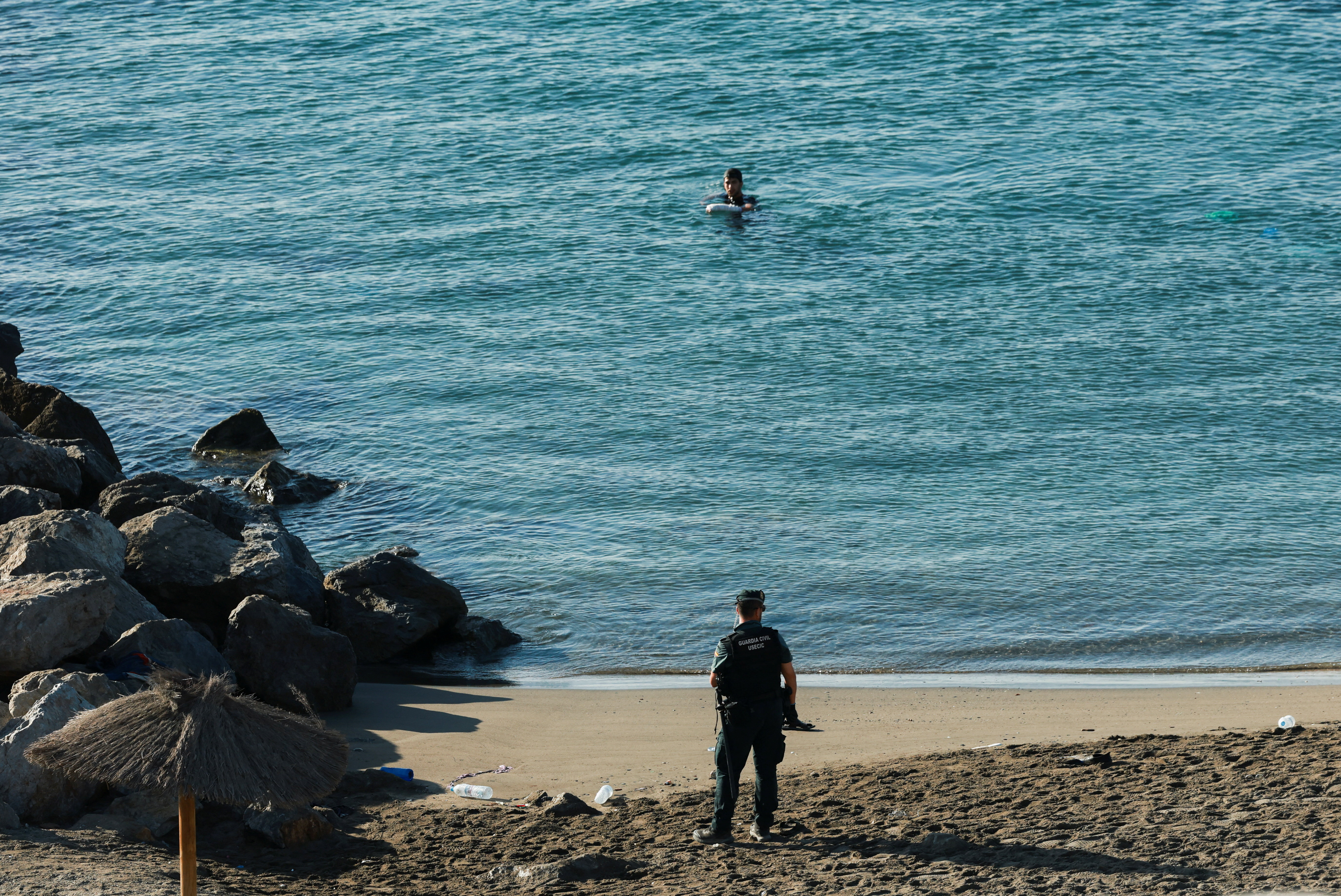 Un miembro de la Guardia Civil permanece en la orilla mientras un migrante marroquí nada para llegar a la playa de El Tarajal de Ceuta.