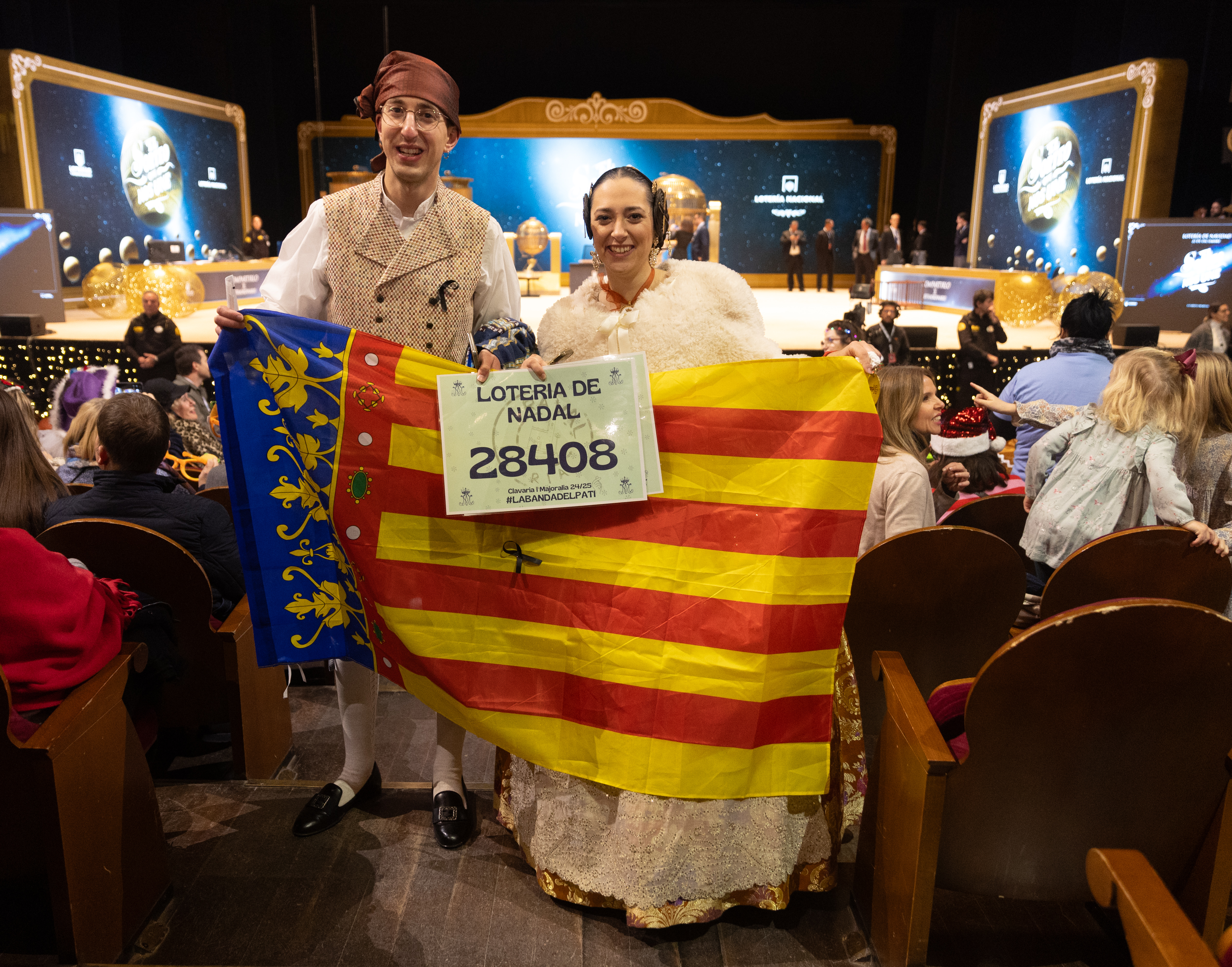 Una pareja vestidos con el traje de fallera y una bandera de la Comunidad Valenciana durante la celebración del Sorteo Extraordinario de la Lotería de Navidad 2024, en el Teatro Real.