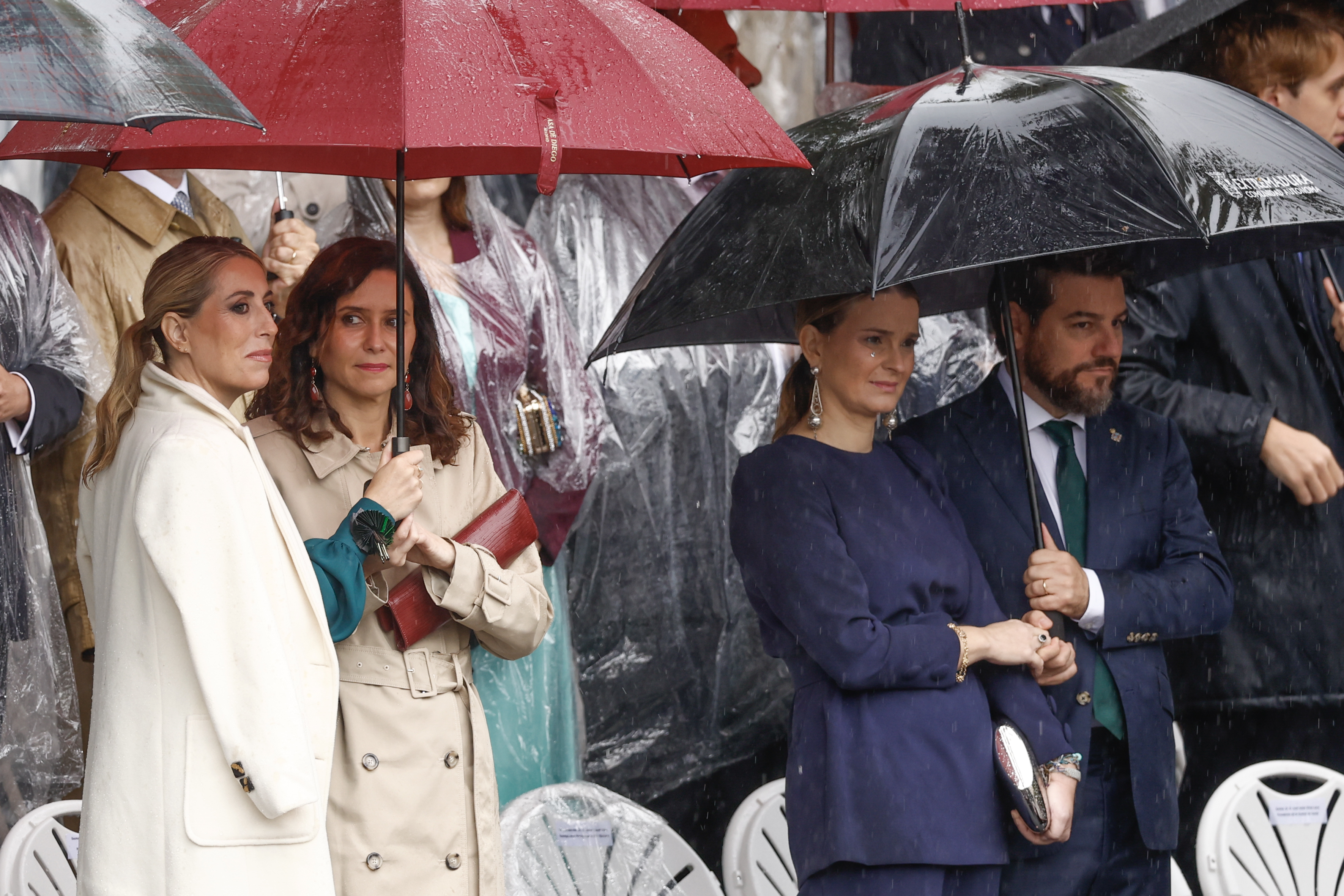 MADRID, 12/10/2024.- La presidenta de la Junta de Extremadura, María Guardiola, la presidenta de la Comunidad de Madrid, Isabel Díaz Ayuso; la presidenta de Baleares, Marga Prohens (2d), y su marido Javier Bonet (d), se protegen de la lluvia durante el desfile del Día de la Fiesta Nacional por el Paseo del Prado de Madrid. EFE/Daniel González
